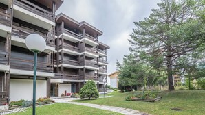 A multi-story residential building with balconies, a paved pathway, and a well-maintained garden with a large tree and flower beds.