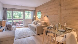 A modern room with a glass dining table set for two, a sofa, and a large window with curtains.