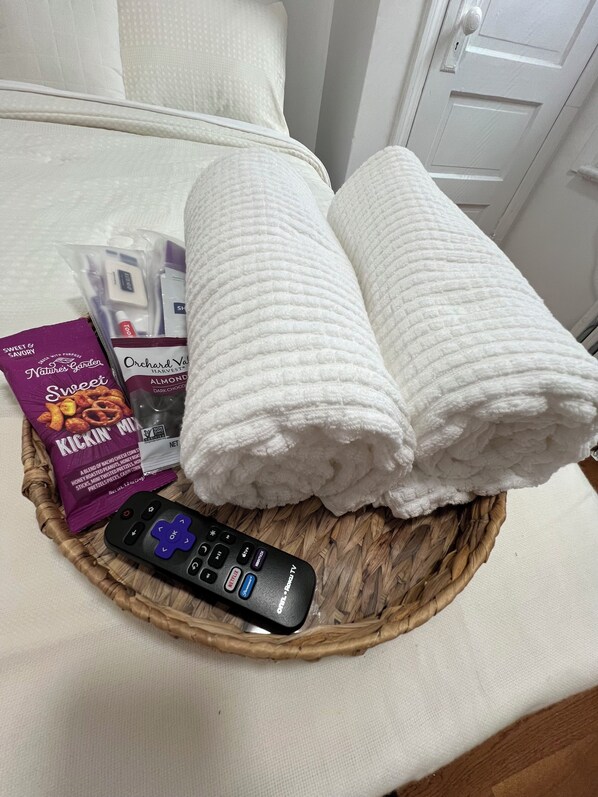 A basket containing a remote control, a package of KICKIN' MID snacks, a package of Orchard Vanilla Almond Dark Chocolate, and two rolled towels on a bed.