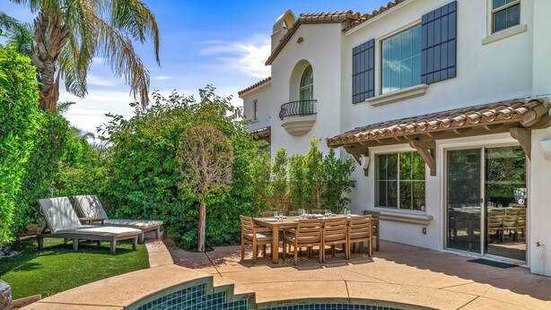 Una piscina con borde curvo, un patio con mesa y sillas, y una casa con balcón.