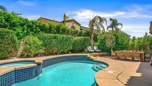 Una piscina con borde de azulejos, rodeada de vegetación y un patio con mobiliario exterior.