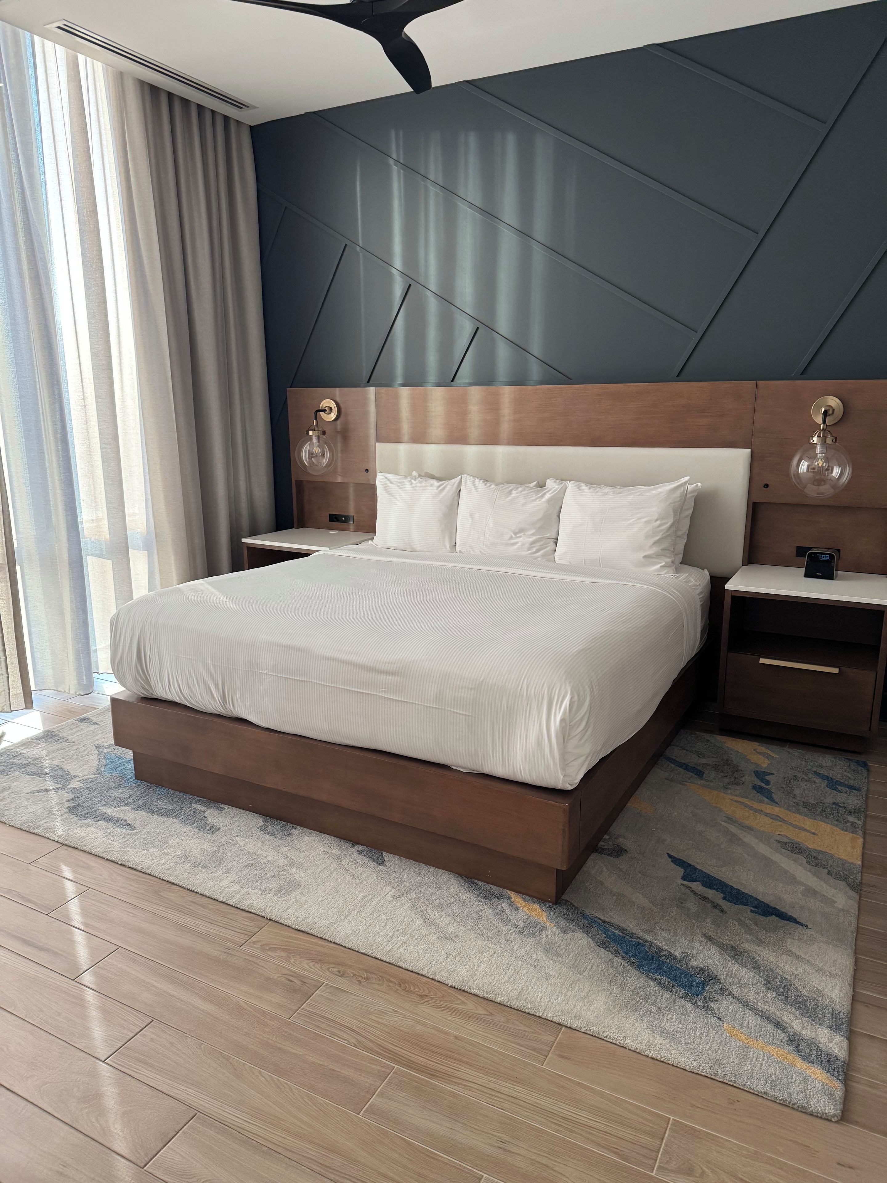 A modern hotel room with a large bed, wooden headboard, bedside tables, and a view of the outdoors.