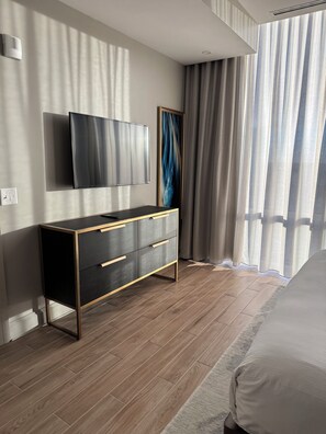 Quarto moderno com piso de madeira, cômoda preta e dourada, televisão de tela plana e uma grande janela com cortinas.