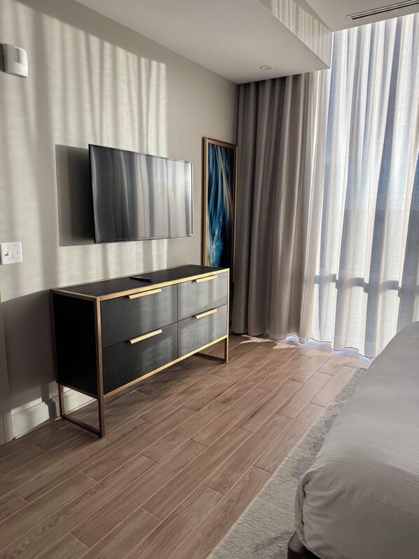 Een moderne slaapkamer met een houten vloer, een zwart en gouden dressoir, een flatscreen televisie en een groot raam met gordijnen.