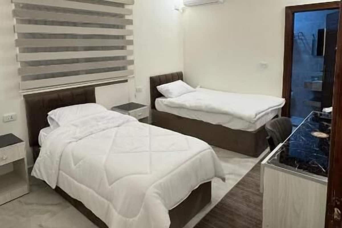 A bedroom with two beds, a desk, a mirror, and a window with blinds.