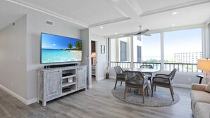A modern living room with a large TV, a dining area, and a balcony with a view of the city. - Luxurious 2BR Gulfview Condo On Siesta Key Steps To The Beach Gulf Horizon (Sarasota)