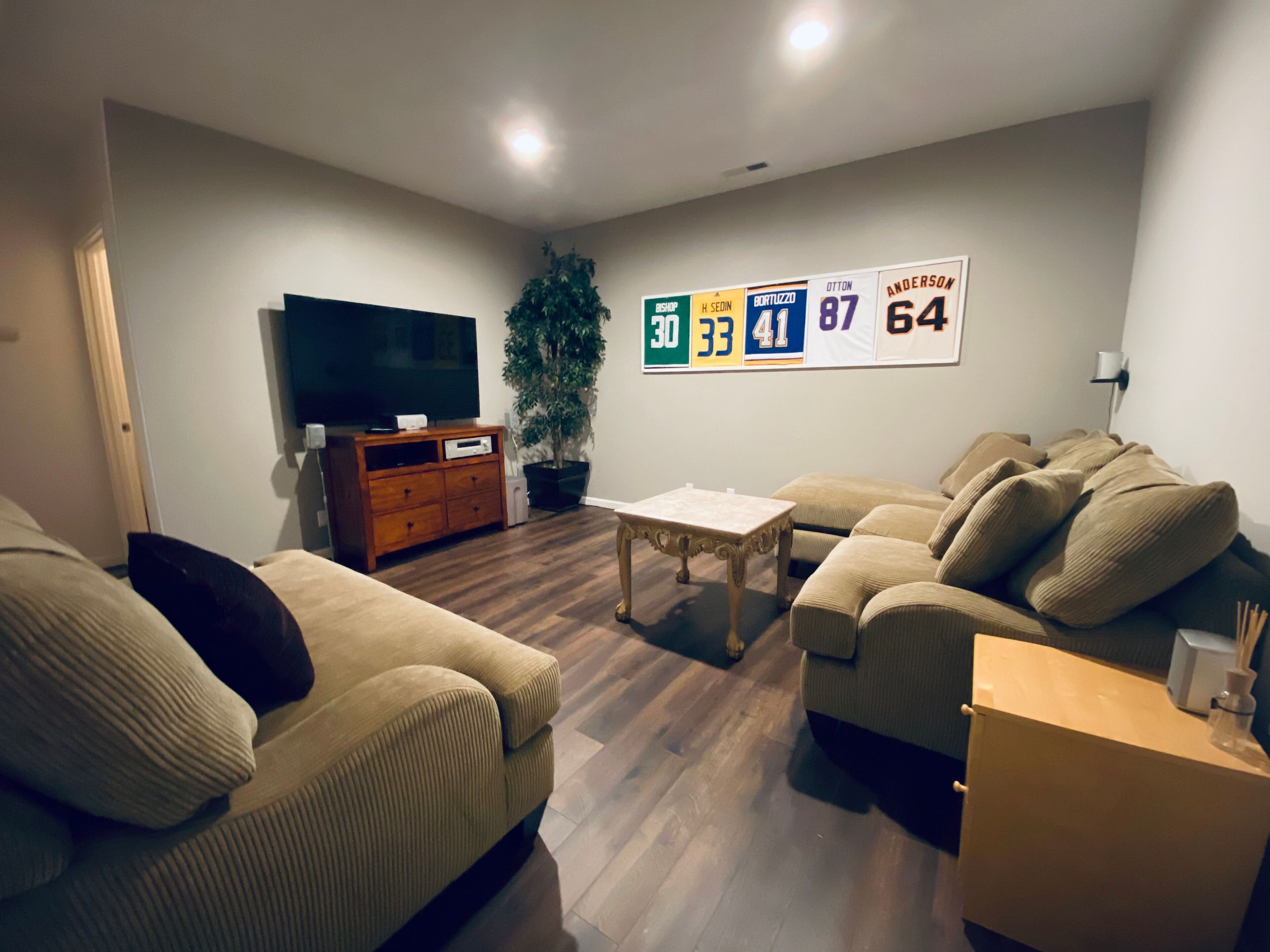 Una sala de estar con un conjunto de sofá, una mesa de centro, un televisor y una pared decorada con camisetas deportivas.