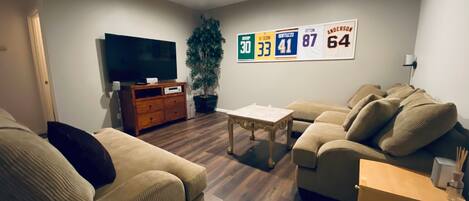 Una sala de estar con un conjunto de sofá, una mesa de centro, un televisor y una pared decorada con camisetas deportivas.