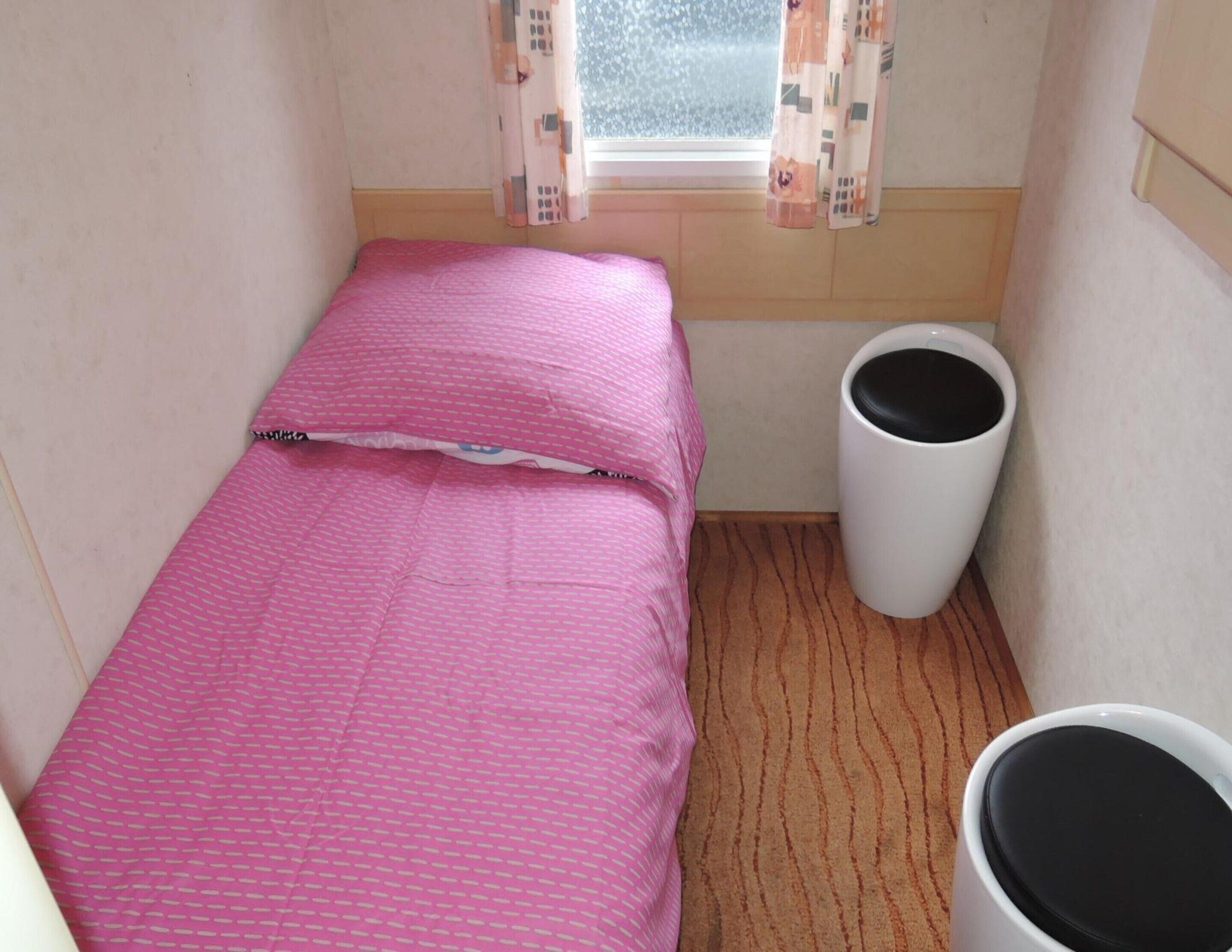 Ein kleines Zimmer mit einem Einzelbett, einer rosa Tagesdecke, einer weißen Mülltonne und einem Fenster mit gemusterten Vorhängen.