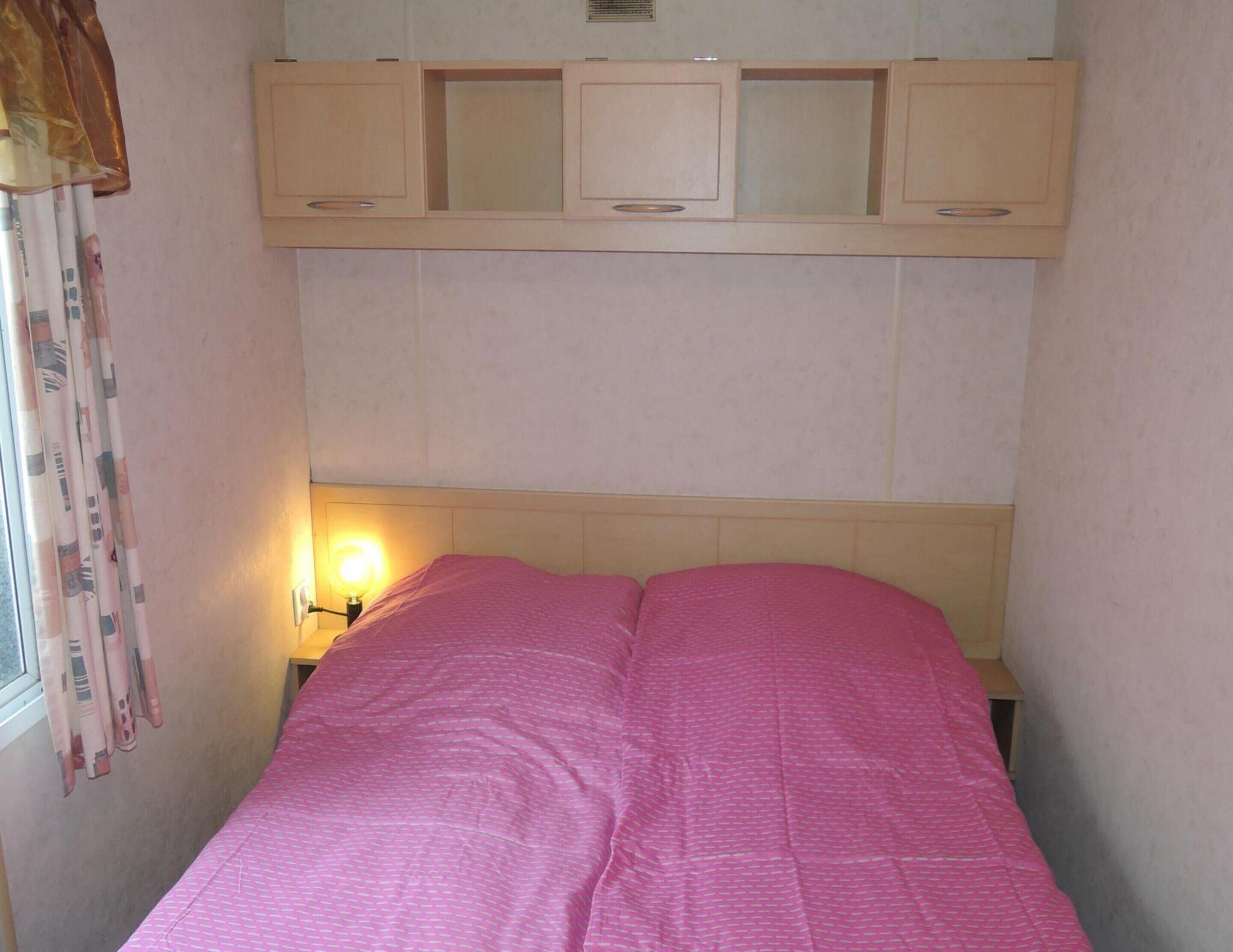 Una habitación pequeña y estrecha con una cama individual, una mesita de noche con lámpara y una ventana con cortinas.