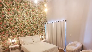 A bedroom with a floral wallpaper, a bed with white bedding, two bedside tables, a window with curtains, and a fluffy chair. - Charming 3-bedroom house with WiFi, AC, Pool, Lake View, by Disney. (Kissimmee)