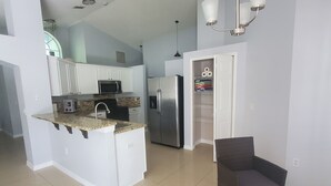 A modern kitchen with white cabinets, a stainless steel refrigerator, and a built-in microwave. There is a dining area with wicker chairs and a glass table. - Charming 3-bedroom house with WiFi, AC, Pool, Lake View, by Disney. (Kissimmee)