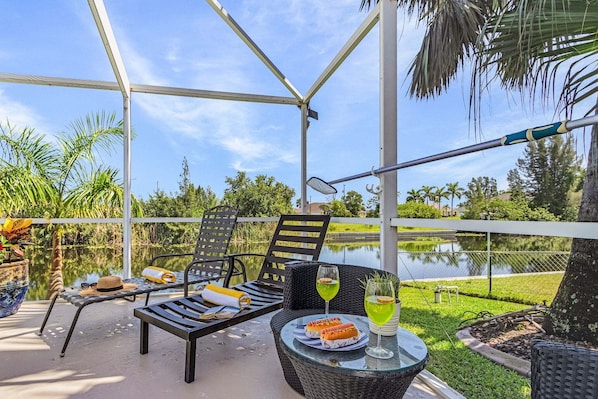 Un patio al aire libre con un área de estar cubierta, una mesa con comida y bebidas, y vistas a un cuerpo de agua y follaje.