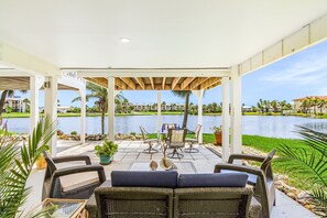 A covered patio with wicker furniture, a table, and chairs, overlooking a lake and buildings.