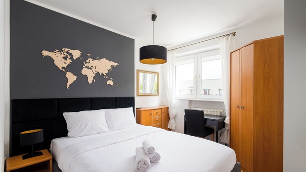 Un lit bien fait, avec du linge de lit blanc et deux serviettes enroulées, une carte du monde accrochée au mur, une armoire en bois et un bureau avec une chaise.