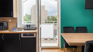 Une cuisine moderne équipée d’un lave-vaisselle, d’une machine à café et d’un évier. Un coin repas est aménagé avec une table et des chaises. Une fenêtre donne sur un balcon d’où l’on peut admirer les bâtiments et les arbres.