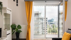 Un salon moderne avec une porte vitrée donnant sur un balcon, une plante en pot et un canapé gris agrémenté d’un coussin jaune.