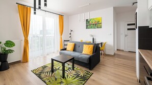 Un salon moderne avec un canapé gris, des coussins jaunes, une table basse noire et un tapis à motifs verts. On y trouve également un coin repas avec une petite table et des chaises, ainsi qu’un coin cuisine doté d’un plan de travail noir.