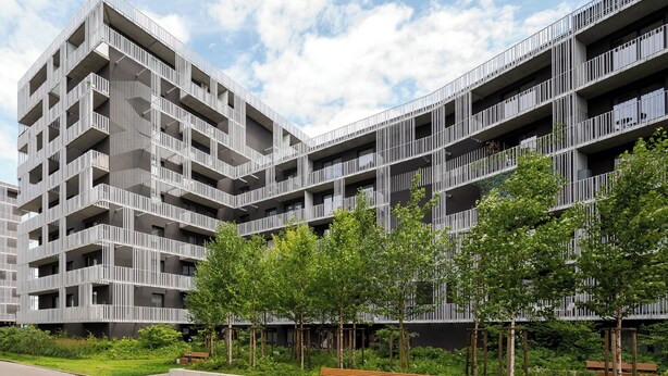 Un immeuble moderne à plusieurs étages, doté de balcons et agrémenté d’un espace paysager avec des arbres et des bancs.