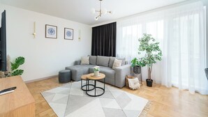 A modern living room with a grey sofa, a small round table with a plant, a large window with sheer curtains, and a wooden floor. - Wrzeciono Comfortline Apartment (Warszawa)