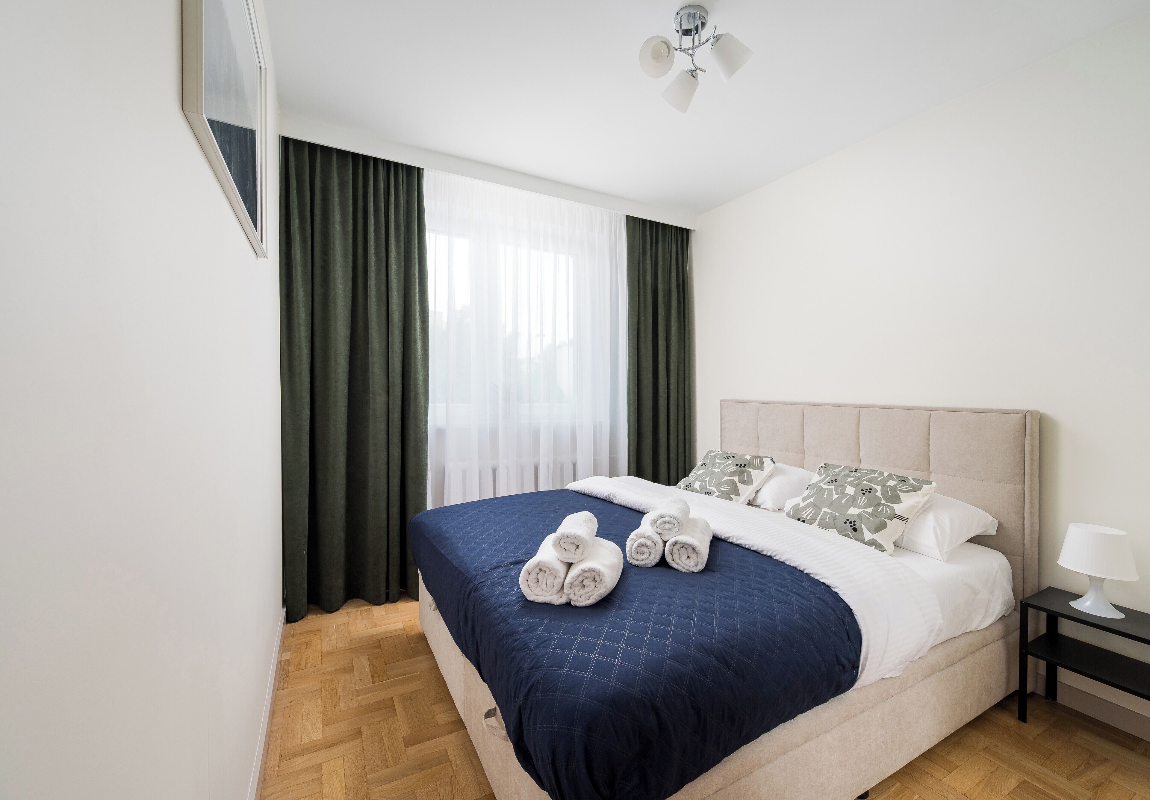 A neatly made bed with a blue bedspread, white pillows, and rolled towels. There is a nightstand with a lamp, and a ceiling fan above.