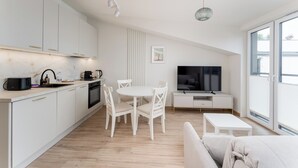 A modern kitchen with white cabinets, a dining area with a round table and chairs, a TV on a white console, and a large window offering a view. - Kalwaryjska Elegant Stay Studio 18B (Kraków)