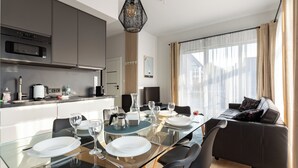 Eine moderne Küche mit dunklen Schränken, einer Mikrowelle und einer Kaffeemaschine. Ein Esstisch mit Stühlen und einer Glasplatte ist gedeckt mit Tellern, Gläsern und Besteck. Im Wohnbereich steht ein schwarzes Sofa mit Blick ins Freie durch große Fenster.