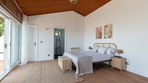 Un dormitorio con cama, mesitas de noche, un banco, una puerta y vistas al exterior.