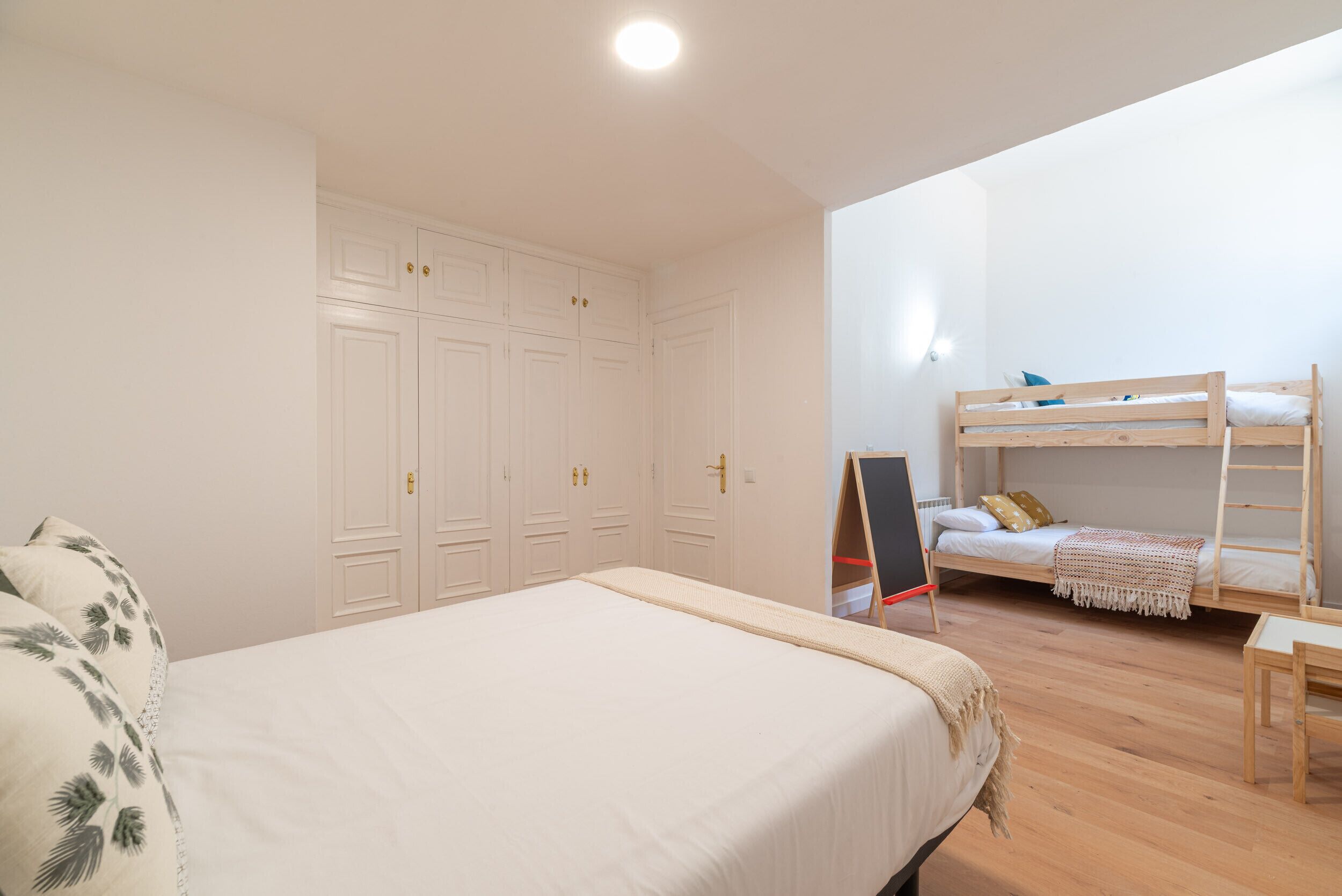 A bedroom with a large bed, a wooden bunk bed, white wardrobes, and a small table.