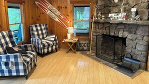 Ein gemütliches Lodge-Interieur mit Holzwänden, einem Kamin aus Stein, karierten Sesseln und einer Wanddekoration zum Thema Skifahren.