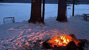 Schneebedeckte Landschaft mit Lagerfeuer, Bäumen und Picknicktisch.