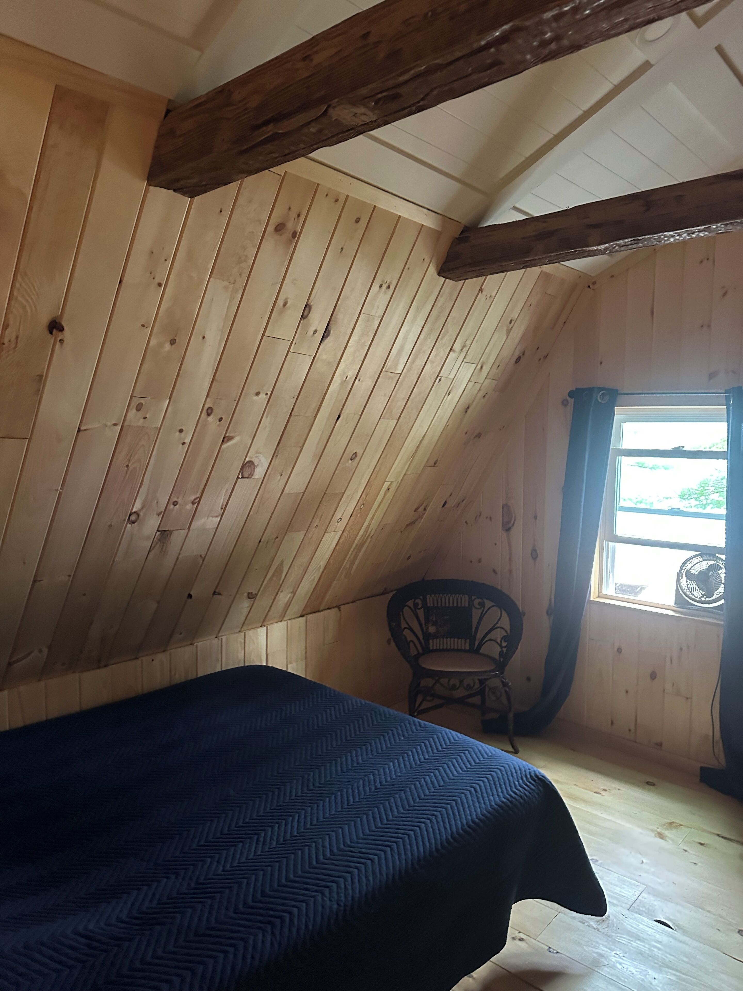 A room with a sloped ceiling, wooden walls, a bed with a dark patterned bedspread, a chair, and a window with curtains.