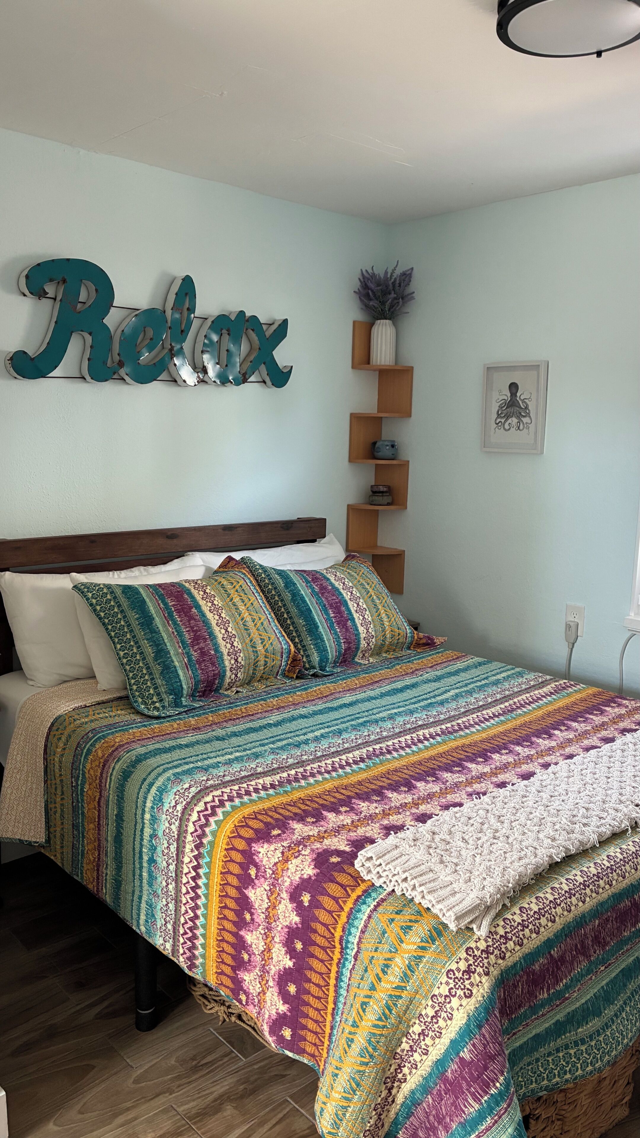 A bedroom with a bed, a wall-mounted shelf with a vase and a framed picture, and a wall decoration spelling 'Relax'.