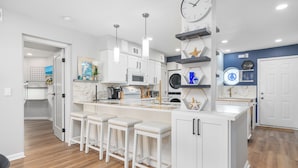 Una cocina moderna con armarios blancos, una isla central con taburetes, microondas, horno y fregadero. Hay objetos decorativos en las repisas y un reloj en la pared.