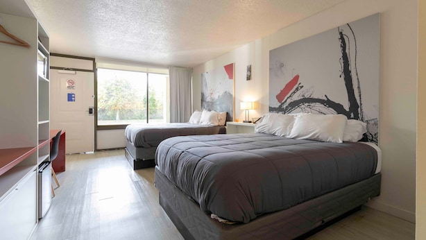 A hotel room with two beds, a desk, and a chair. There is a large abstract painting on the wall and a window providing natural light. - Magical Studio for 4 Shuttle to Disney (Kissimmee)