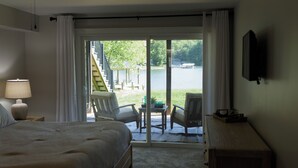 Un dormitorio con cama, mesita de noche, lámpara, televisor y vistas a una terraza con sillas y una mesa que da a un cuerpo de agua.