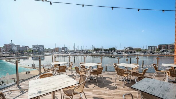 Une terrasse extérieure avec des tables et des chaises en osier, donnant sur une marina où l’on aperçoit des bateaux et des bâtiments en arrière-plan.