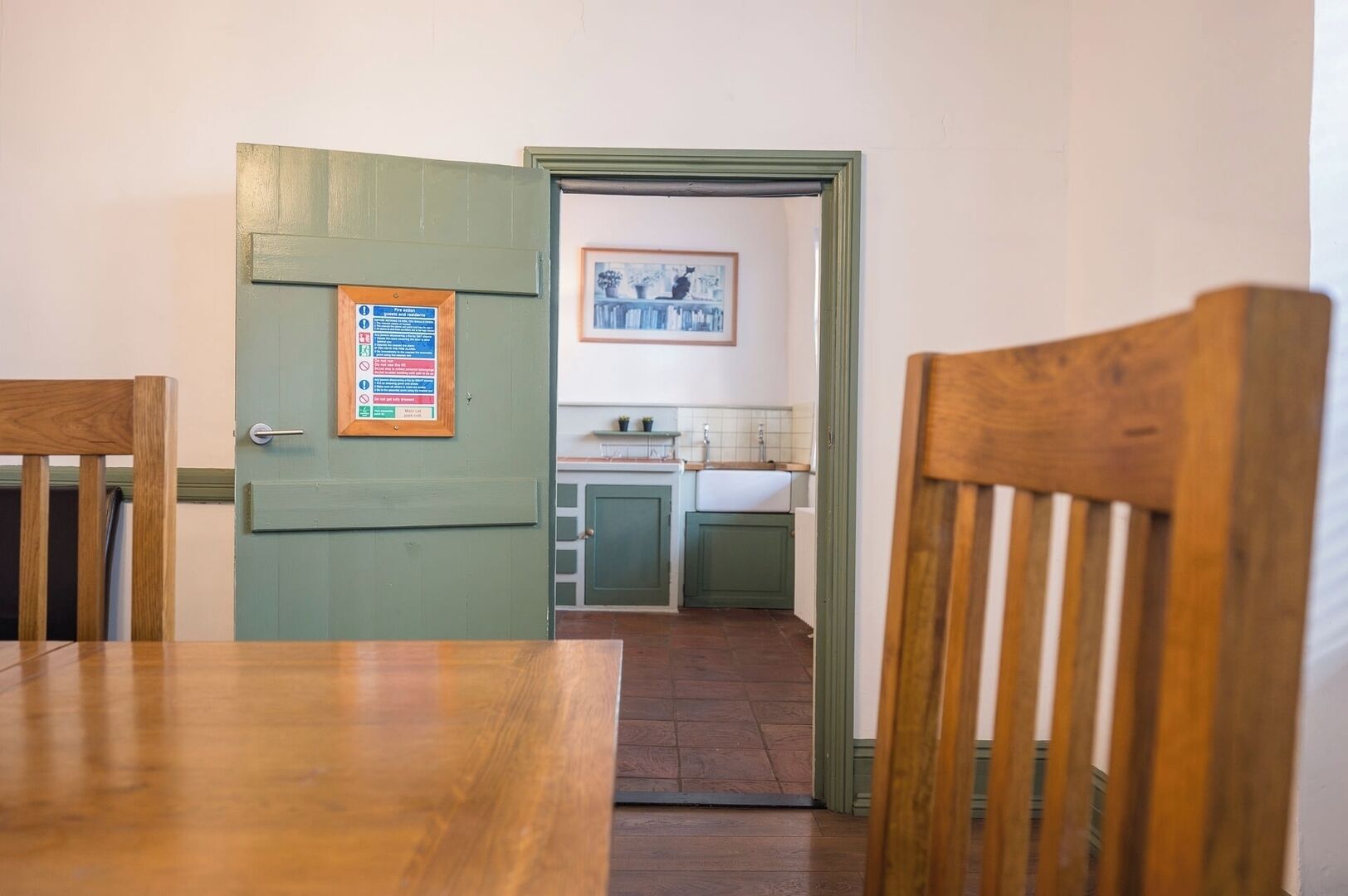 Un comedor con mesa y sillas de madera, una puerta verde con un aviso y vistas a una cocina con pileta y alacenas.