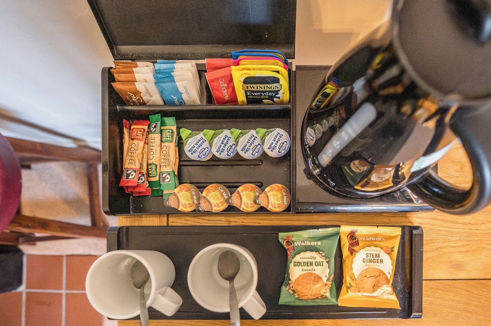 A coffee station with various coffee pods, a coffee maker, two cups, and snacks.
