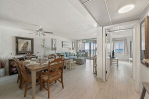 A spacious living area with a dining table set for four, a sofa, and a view of the ocean through large windows.