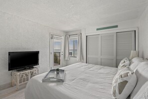 A bedroom with a bed, a television on a stand, a window with curtains, and a door with louvered shutters.