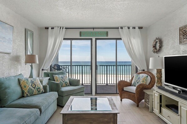 A living room with a sofa, armchair, and coffee table, a television, and a view of the beach through a sliding door.