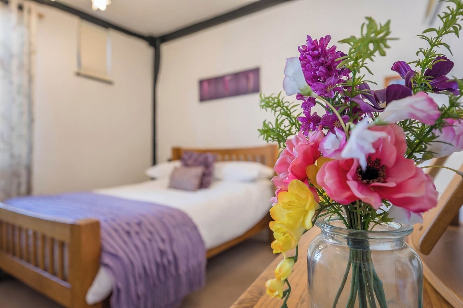Ein Schlafzimmer mit einem hölzernen Bett, einer violetten Decke und einem Blumenstrauß in einer Vase auf einem Nachttisch aus Holz.