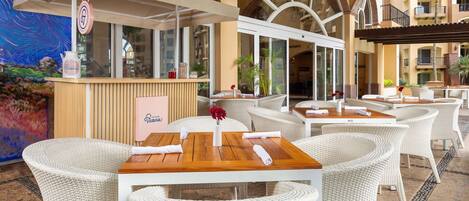 An outdoor seating area with wicker chairs and tables, a bar area with a counter, and a view of the building's interior through glass doors.