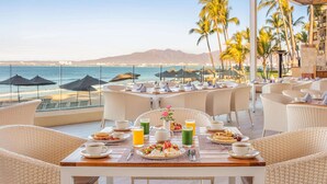 A dining area with wicker furniture, a table set with breakfast, and a view of the ocean and palm trees. - Beachfront Resort w/ Luxurious Amenities-Villa Del Palmar Flamingos Resort & Spa (Bucerias)