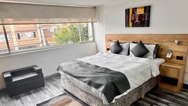 A modern bedroom with a large bed, a wooden headboard, a nightstand, a window with a view of buildings, and a small storage unit. - HOTEL FENIX REAL (Bogotá)