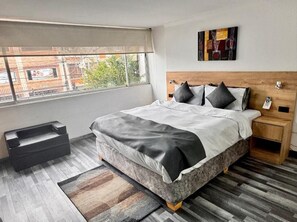 Une chambre moderne avec un grand lit, une tête de lit en bois, une table de chevet, une fenêtre donnant sur des immeubles et un petit meuble de rangement.