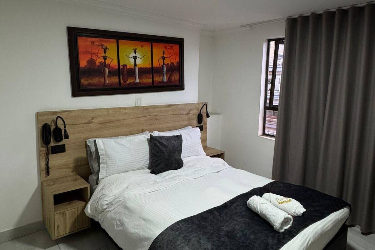 A hotel room with a wooden headboard, a bed with white and dark grey bedding, a nightstand with a phone and a lamp, a painting above the bed, and a window with curtains.