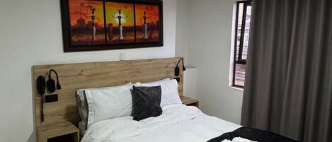 Habitación de hotel con cabecera de madera, cama con ropa de cama blanca y gris oscuro, mesita de noche con teléfono y lámpara, un cuadro sobre la cama y una ventana con cortinas.