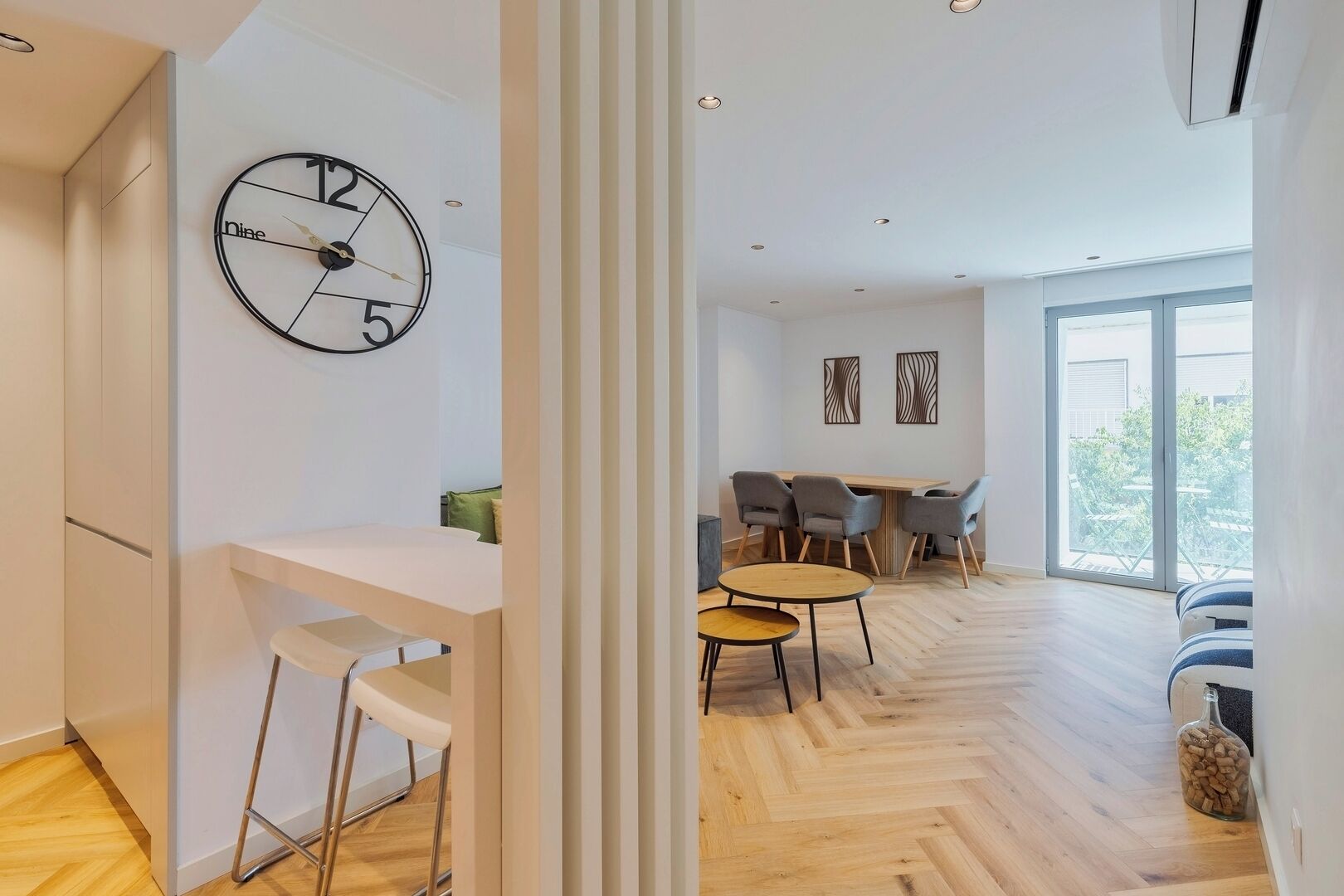 Un salon moderne avec un parquet en bois disposé en chevrons, un coin repas comprenant une table et des chaises en bois, et une grande horloge murale.